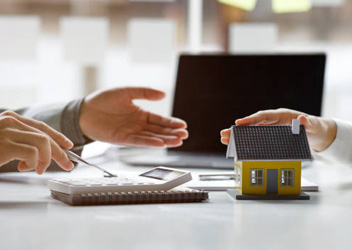 Two hands are seen on a table. One is holding a small toy house, while the other points at a calculator, with a laptop in the background.