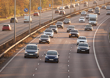 A busy highway with multiple lanes, showing various cars and trucks traveling in both directions. The road is bordered by a concrete barrier and traffic signs, with trees visible in the background under clear skies.
